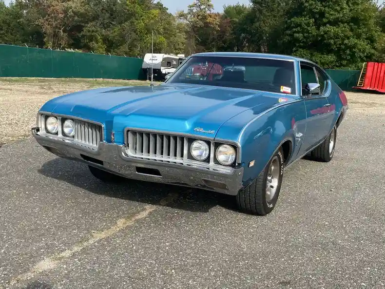 A shiny blue classic American muscle car, likely a 1970s Oldsmobile Cutlass, is parked on asphalt with trees and a green…