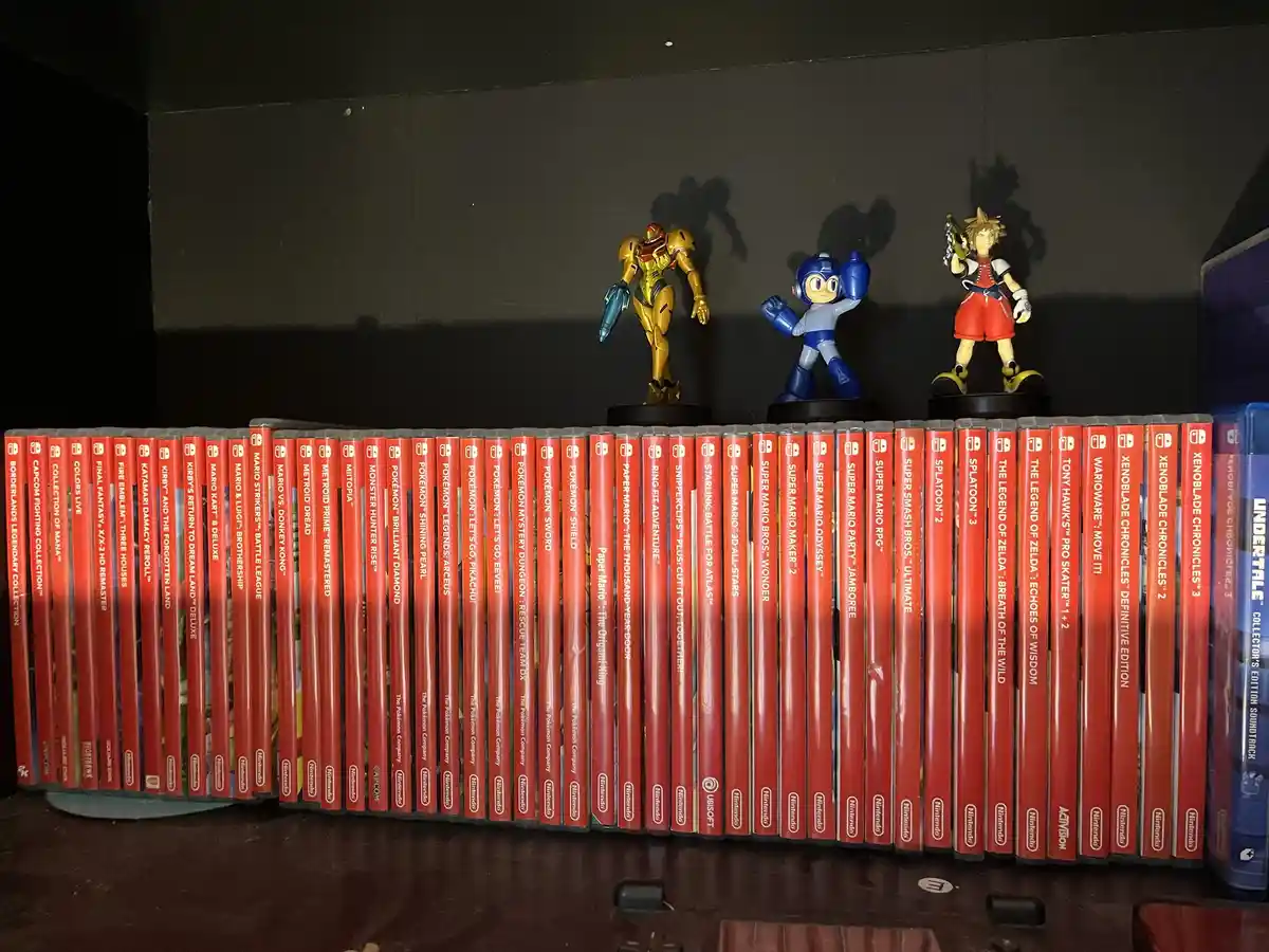 A shelf displays a long row of Nintendo Switch game cases, mostly with red spines, arranged neatly. On top of the cases sit…