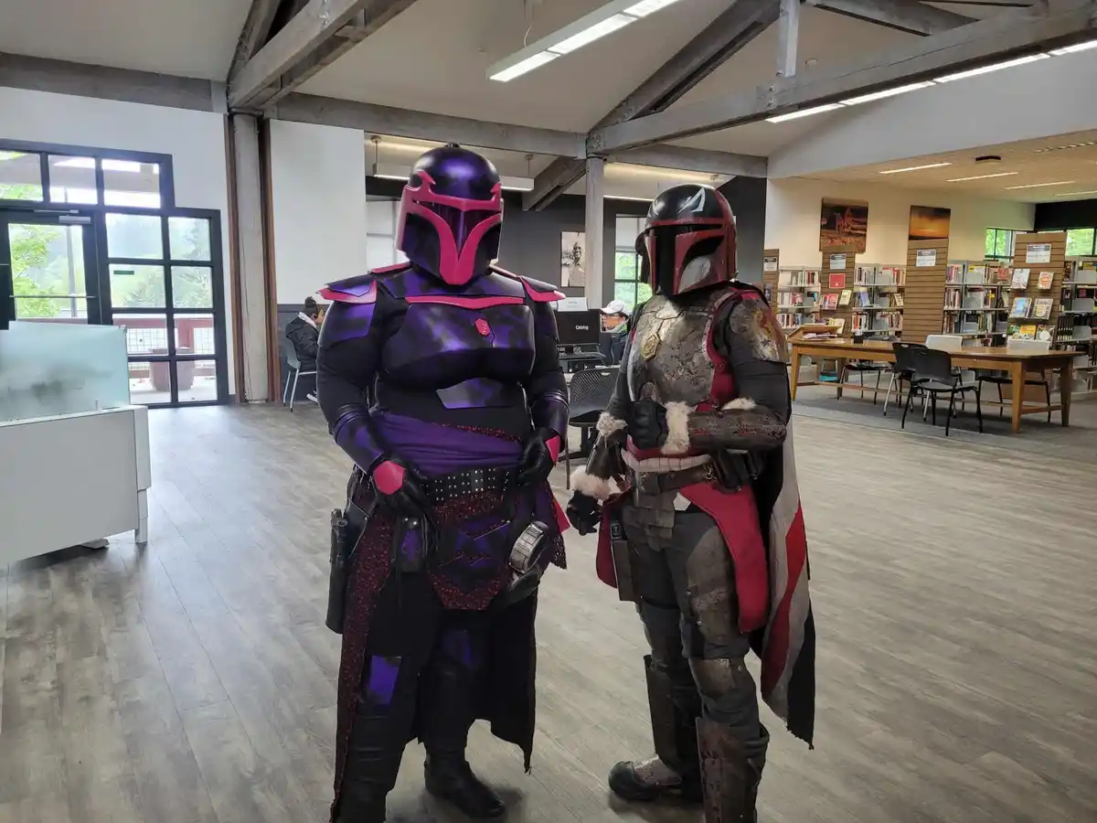 Two people in Mandalorian armor stand inside a modern library, one in purple and pink, the other in weathered red and black…
