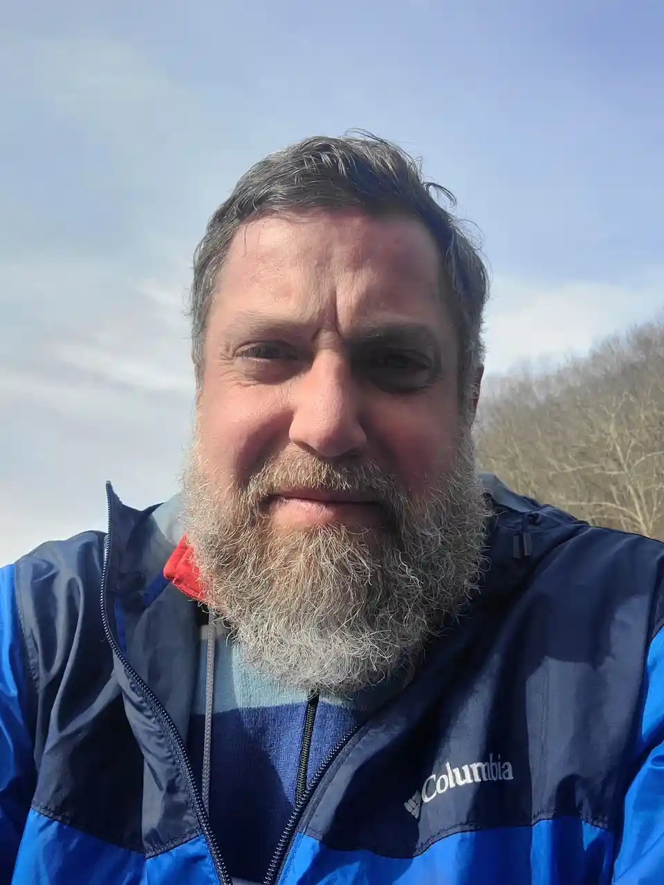 A man with a graying beard and short hair, wearing a blue Columbia jacket over a striped shirt, takes a selfie outdoors…