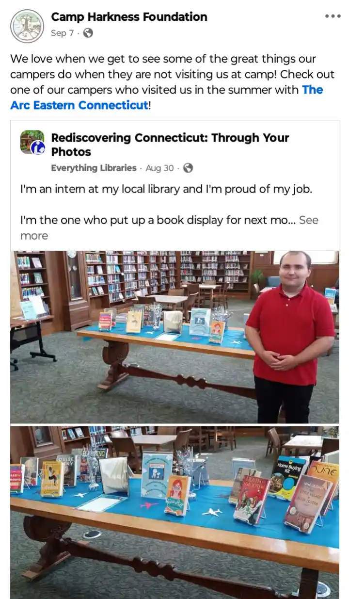 A man in a red polo shirt stands smiling beside a library table covered with a blue cloth and displaying various books in…