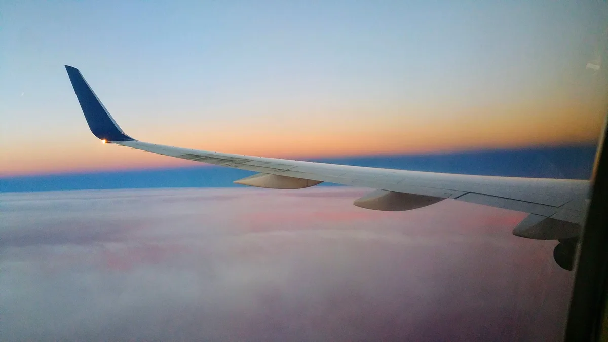 A serene view from an airplane window showing the wing against a colorful sunset sky above clouds. The overlaid text reads…