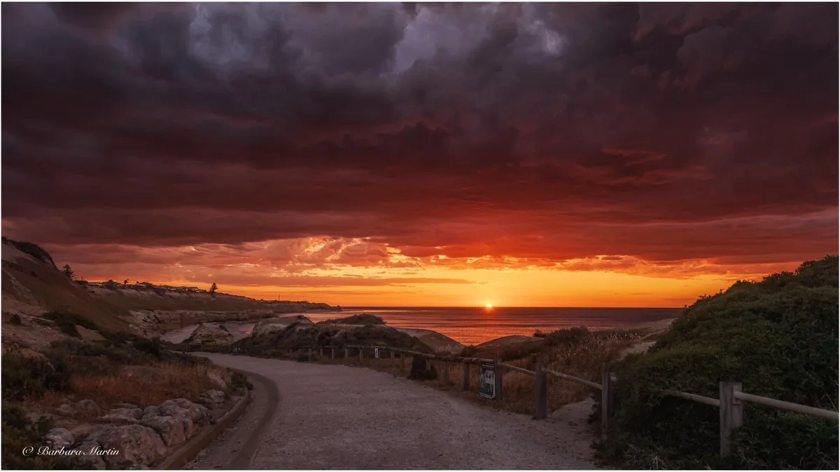 A dramatic sunset photo shows a winding gravel path leading toward the ocean under a sky of fiery orange and ominous dark…