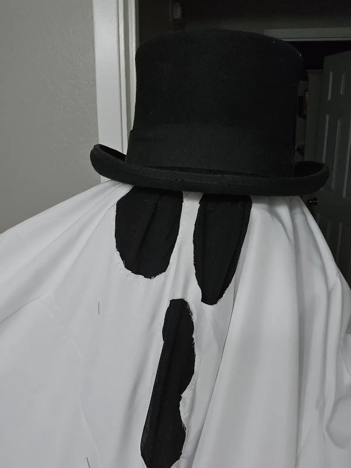 A person wearing a white sheet costume with black fabric cutouts for eyes and a mouth, topped with a black top hat…