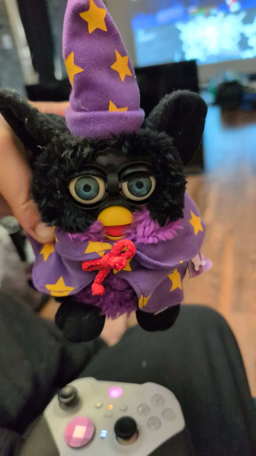 A black, furry Furby toy dressed as a wizard, wearing a purple hat and cape with yellow stars and a red knotted tie, is held…