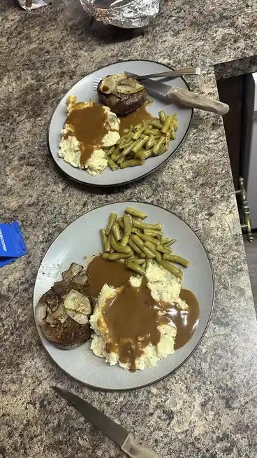 Two plates of food sit on a granite countertop, each with a steak topped with onions, mashed potatoes covered in brown…