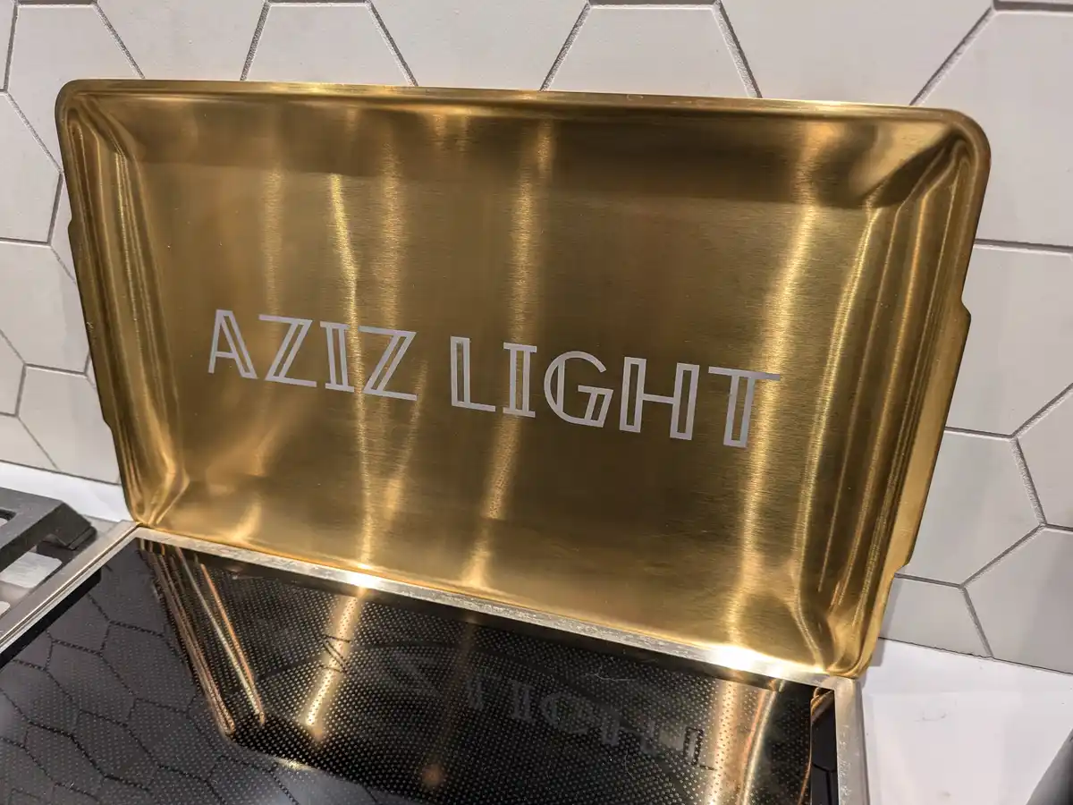 A golden-colored metal baking tray with the words 'AZIZ LIGHT' engraved in white, sits on a black glass stovetop against a…