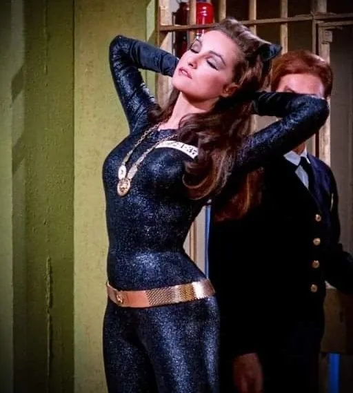 A woman in a sparkly black catsuit with cat ears and a gold belt stands with her hands behind her head, looking smug. Behind…
