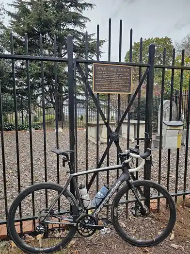 A black Cannondale road bicycle is leaning against a black metal fence at the entrance of the Calvert Family Cemetery, with…