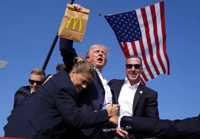 A man in a suit holds up a McDonald's bag while being embraced by another person, with an American flag waving behind them…