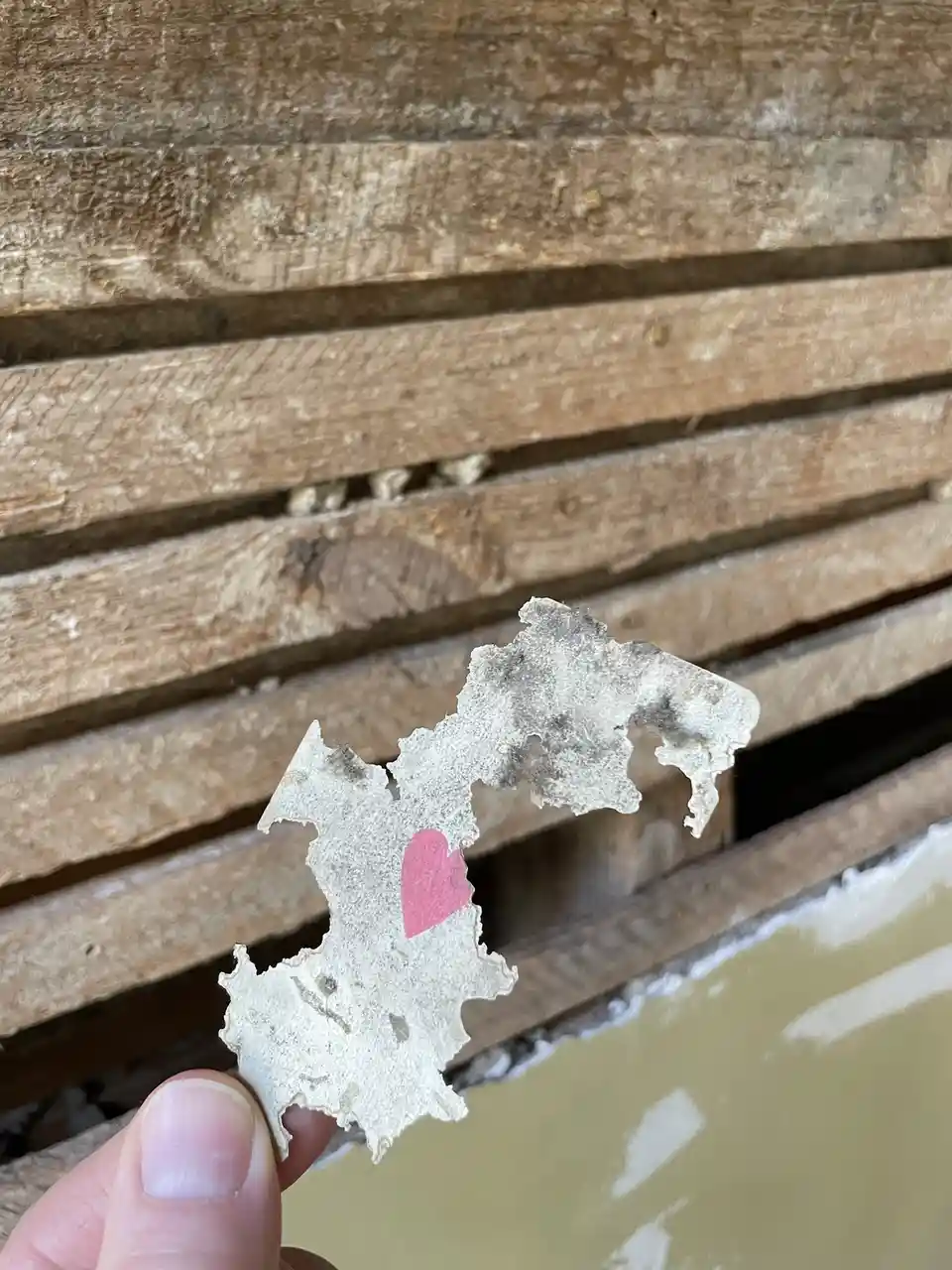 A hand holds up a small, torn, dirty piece of paper with a pink heart drawn on it, against a background of weathered wooden…
