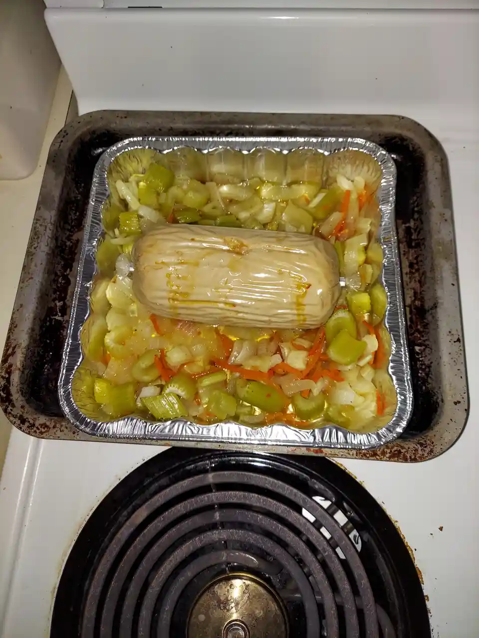A foil tray containing chopped celery, onions, and carrots sits on a metal baking sheet atop a white electric stove burner…