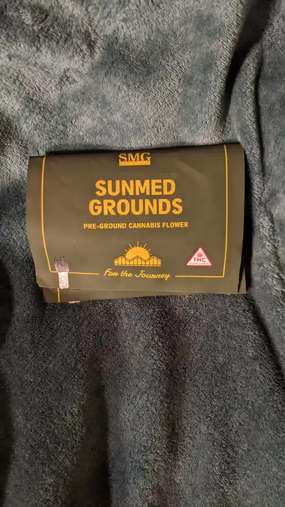 A close-up photo shows a dark green package of 'SMG SUNMED GROUNDS PRE-GROUND CANNABIS FLOWER' resting on a dark gray…