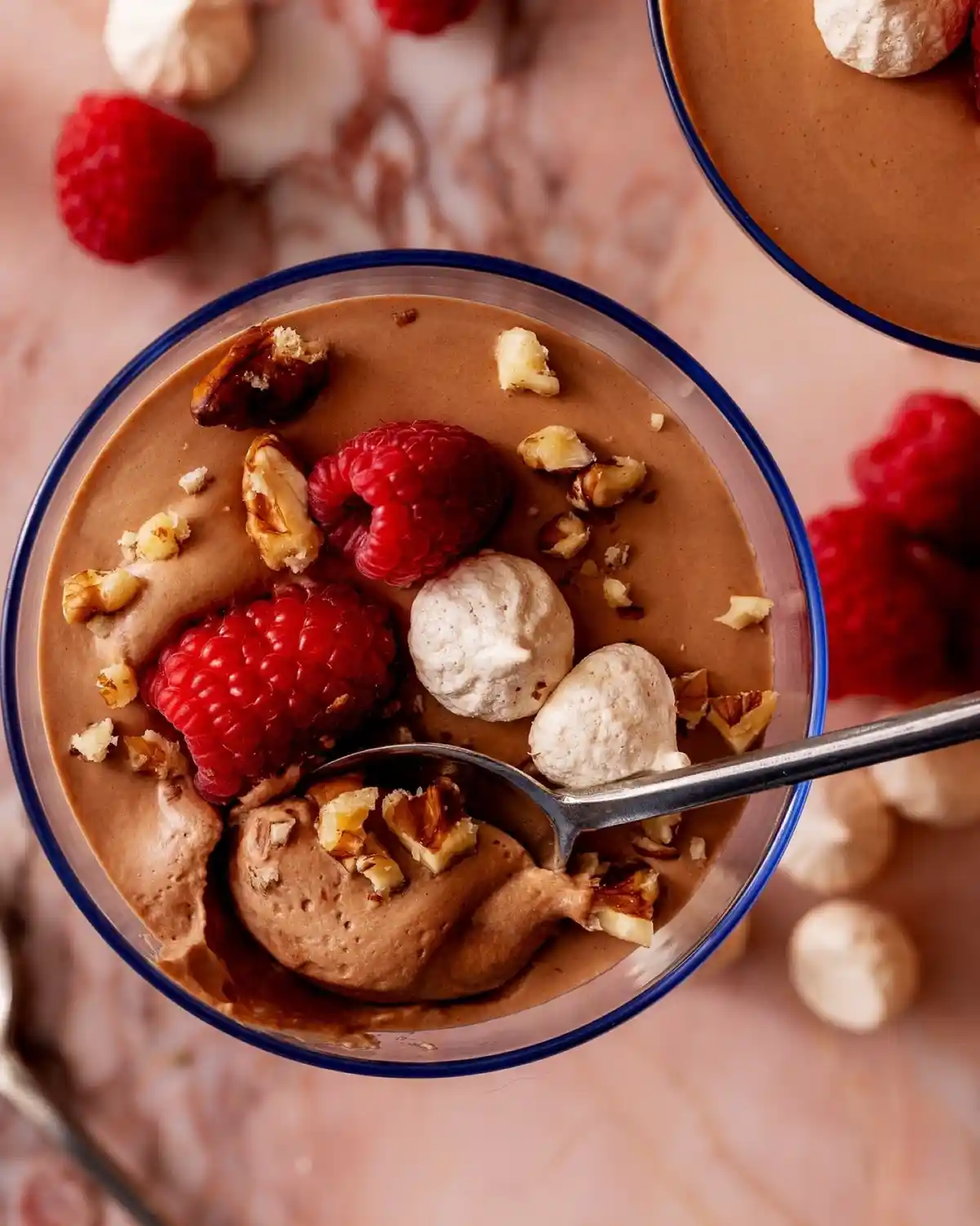 a top-down photo of a glass bowl filled with chocolate pudding, garnished with fresh raspberries, chopped nuts, and small…