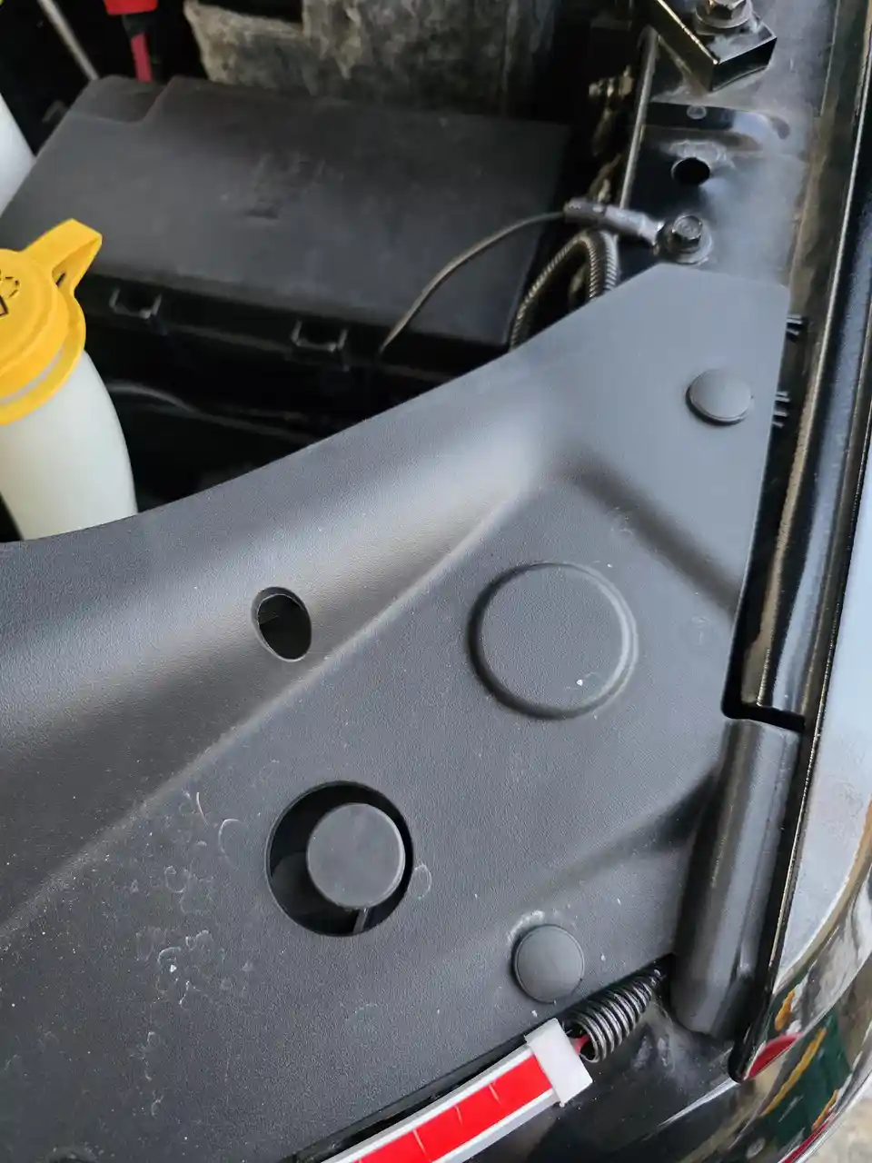 A close-up photo of a black plastic car panel with several circular indentations and holes, next to a yellow-capped fluid…