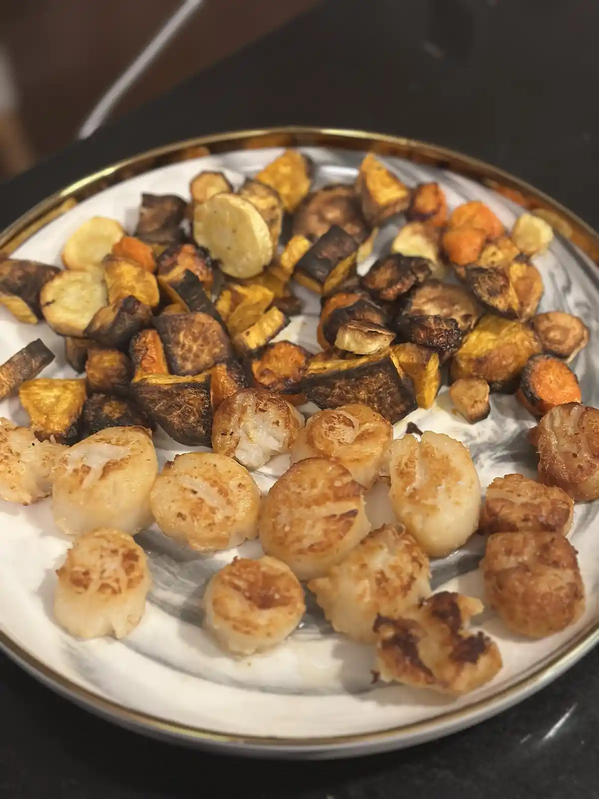 A close-up photo of a white plate with a gold rim, holding a serving of seared scallops and a mix of roasted root vegetables…