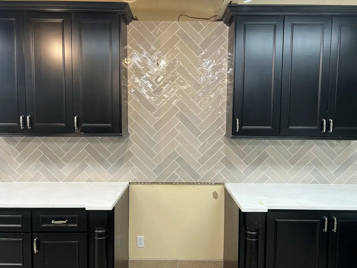 A kitchen backsplash with a herringbone pattern of glossy gray tiles is installed between black cabinets, but a large…