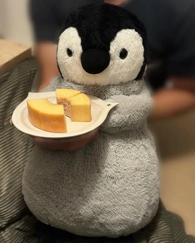 A grey and black plush penguin is holding a small white plate with three slices of yellow cake on it. The cake has a crumbly…