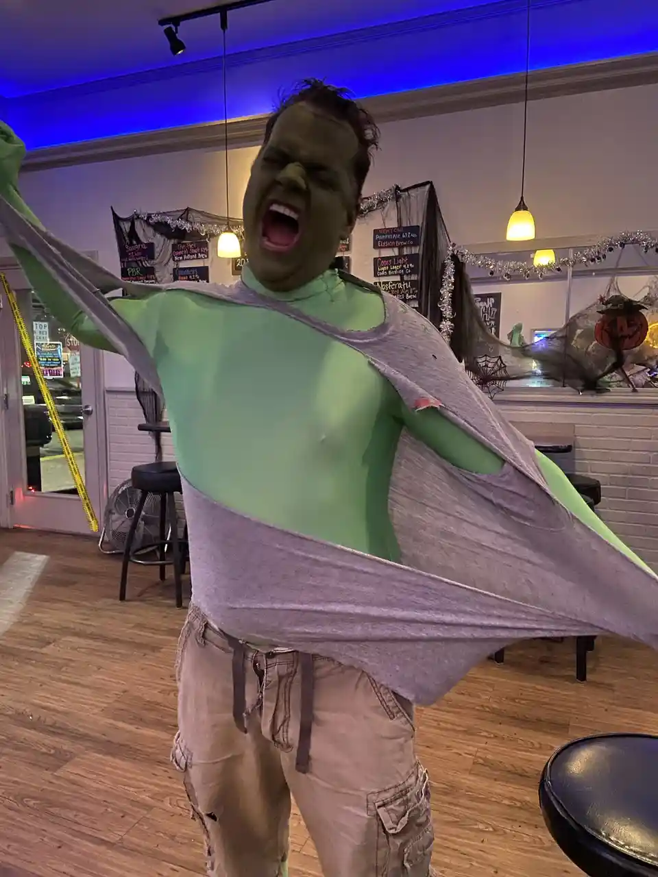 A person with green face paint and a green bodysuit is dramatically ripping off a grey t-shirt in a Halloween-themed bar…