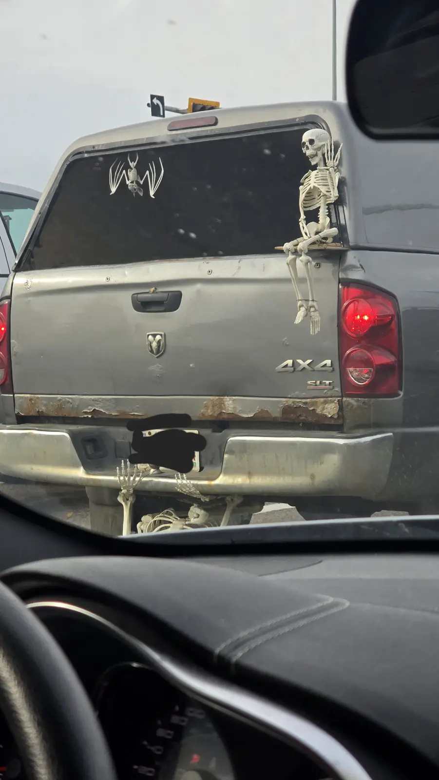A silver Dodge Ram 4x4 pickup truck has multiple plastic skeletons attached to it: one sitting on the tailgate, one hanging…