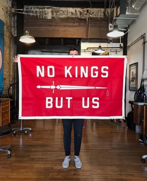A person stands in an industrial-style room holding a large red banner with white text that reads 'NO KINGS BUT US' and a…