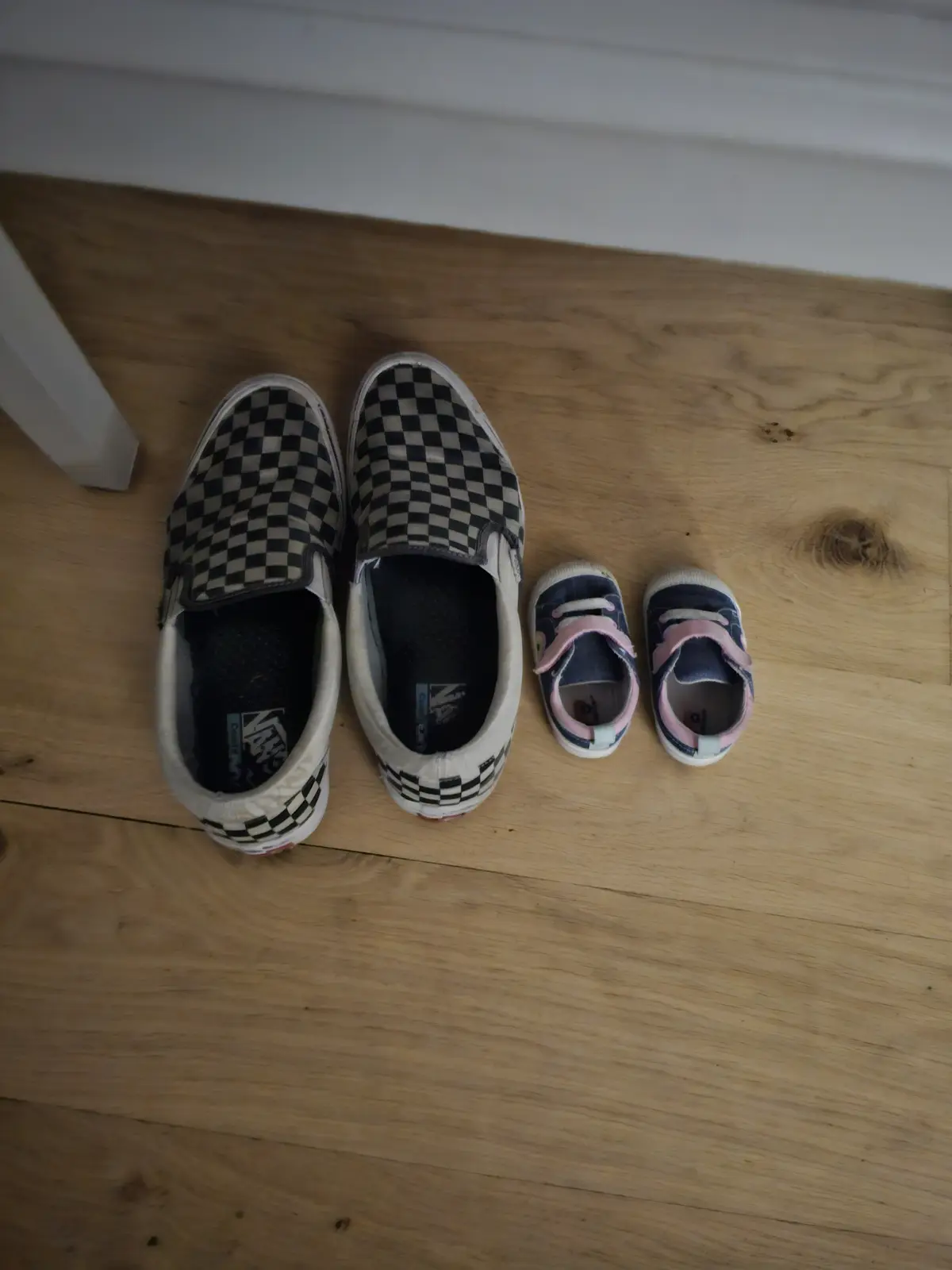 A top-down photo shows a pair of large, worn black-and-white checkered adult slip-on shoes next to a pair of tiny blue baby…