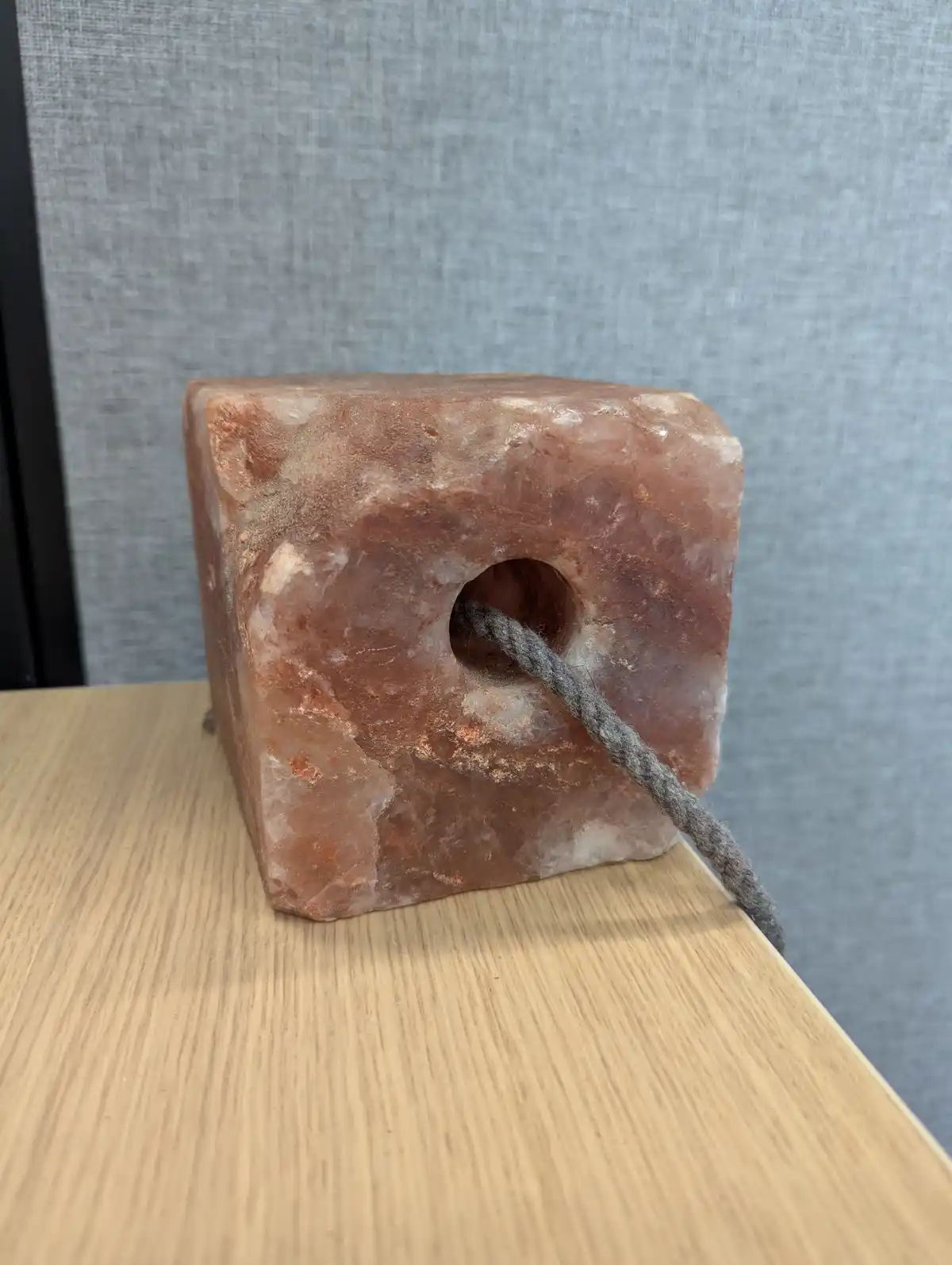 A rough, pinkish-orange salt block with a hole drilled through its center sits on a light wood surface, with a gray rope…