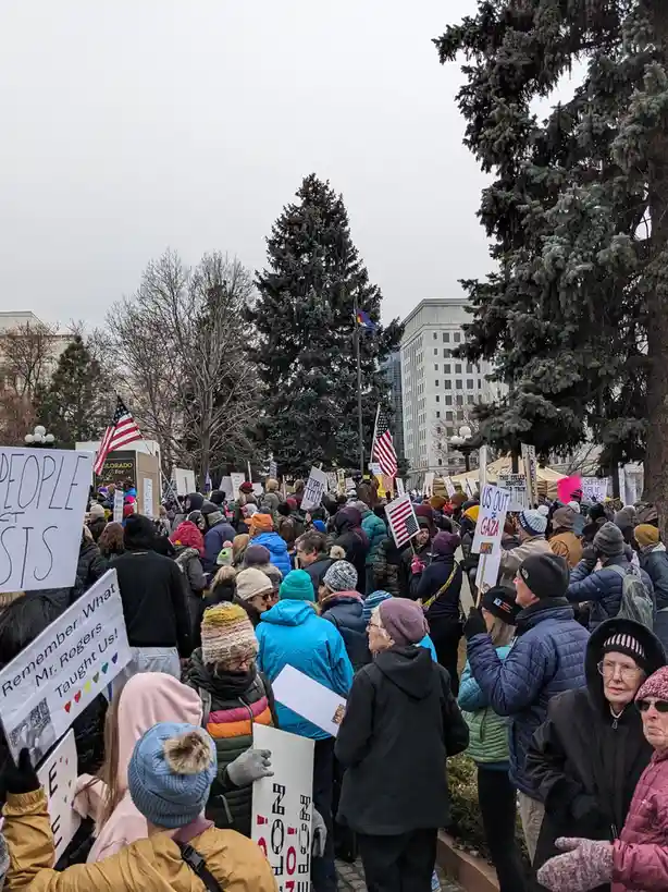 A crowd of people dressed in winter coats and hats are gathered outdoors at a protest, holding signs with messages like 'US…