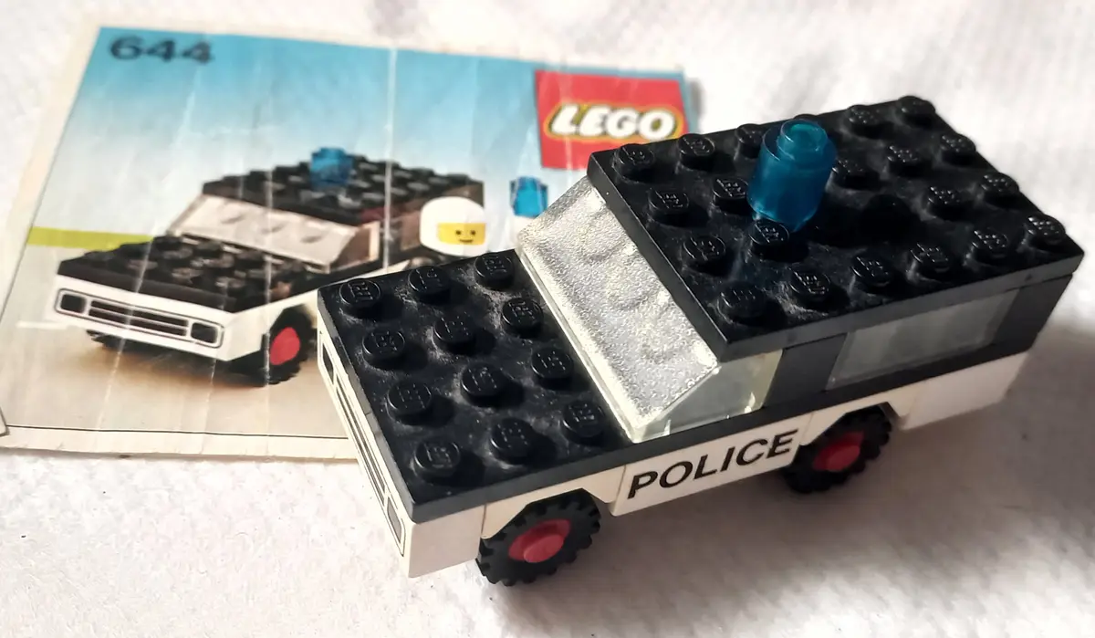 A handmade LEGO police car, constructed from black and white bricks with red wheels and a blue light, sits next to its…
