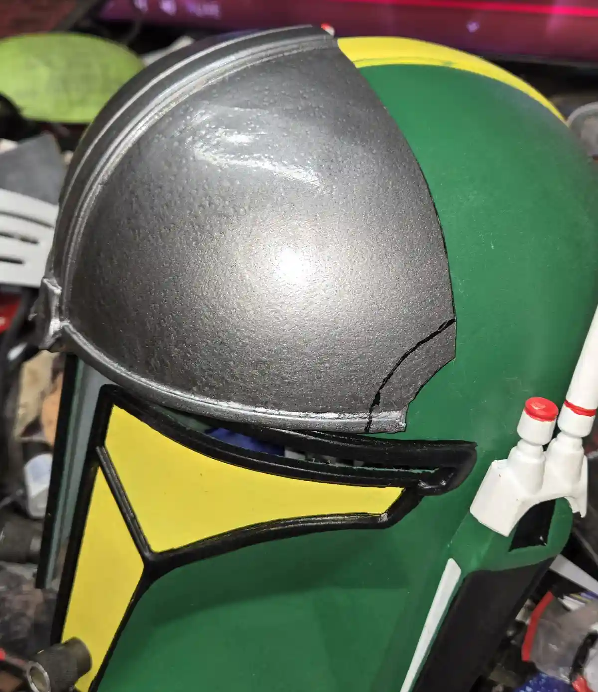 A close-up of a green and yellow Mandalorian-style helmet with a cracked, silver-colored dome section. The text overlay…