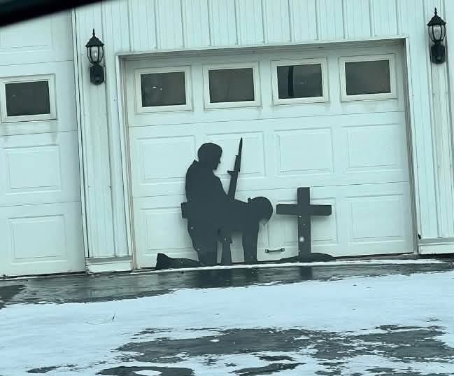 A black silhouette cutout of a soldier kneeling beside a cross is mounted on a white garage door. The soldier holds a rifle…
