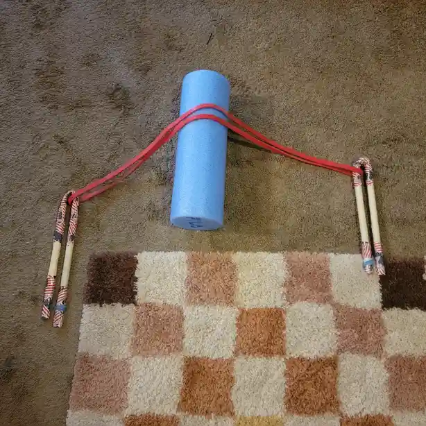 A blue foam roller sits on a brown carpet, with a red resistance band looped around it and attached to two chopsticks…