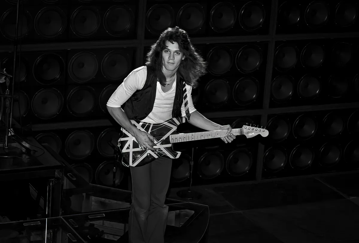 A black-and-white photo shows a young man with long, wavy hair holding an electric guitar with a distinctive white geometric…
