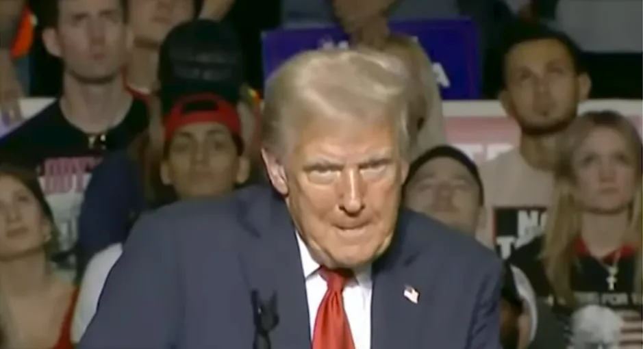A close-up of Donald Trump speaking at a rally, wearing a blue suit and red tie, with a stern expression. Behind him, a…
