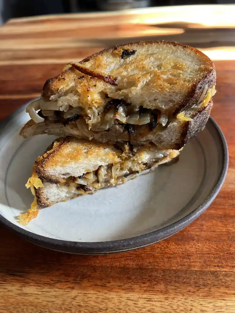 A close-up photo shows a grilled sandwich cut in half and stacked on a ceramic plate, revealing melted cheese and…