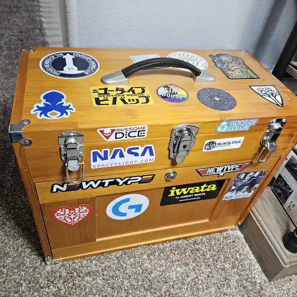 A wooden chest with metal latches and a black handle is covered in numerous stickers, including NASA, Newtype, Iwata…