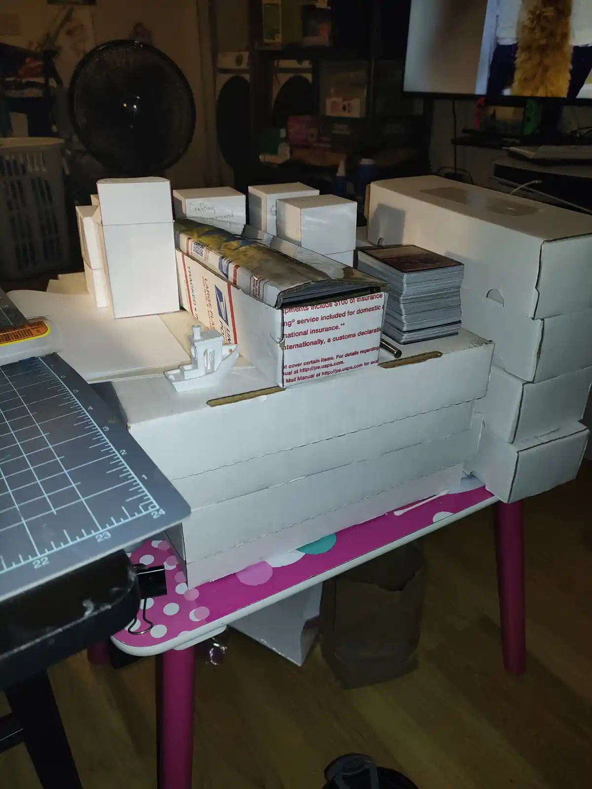 A cluttered crafting table with a stack of white cardboard boxes forming a makeshift table surface, topped with a USPS…