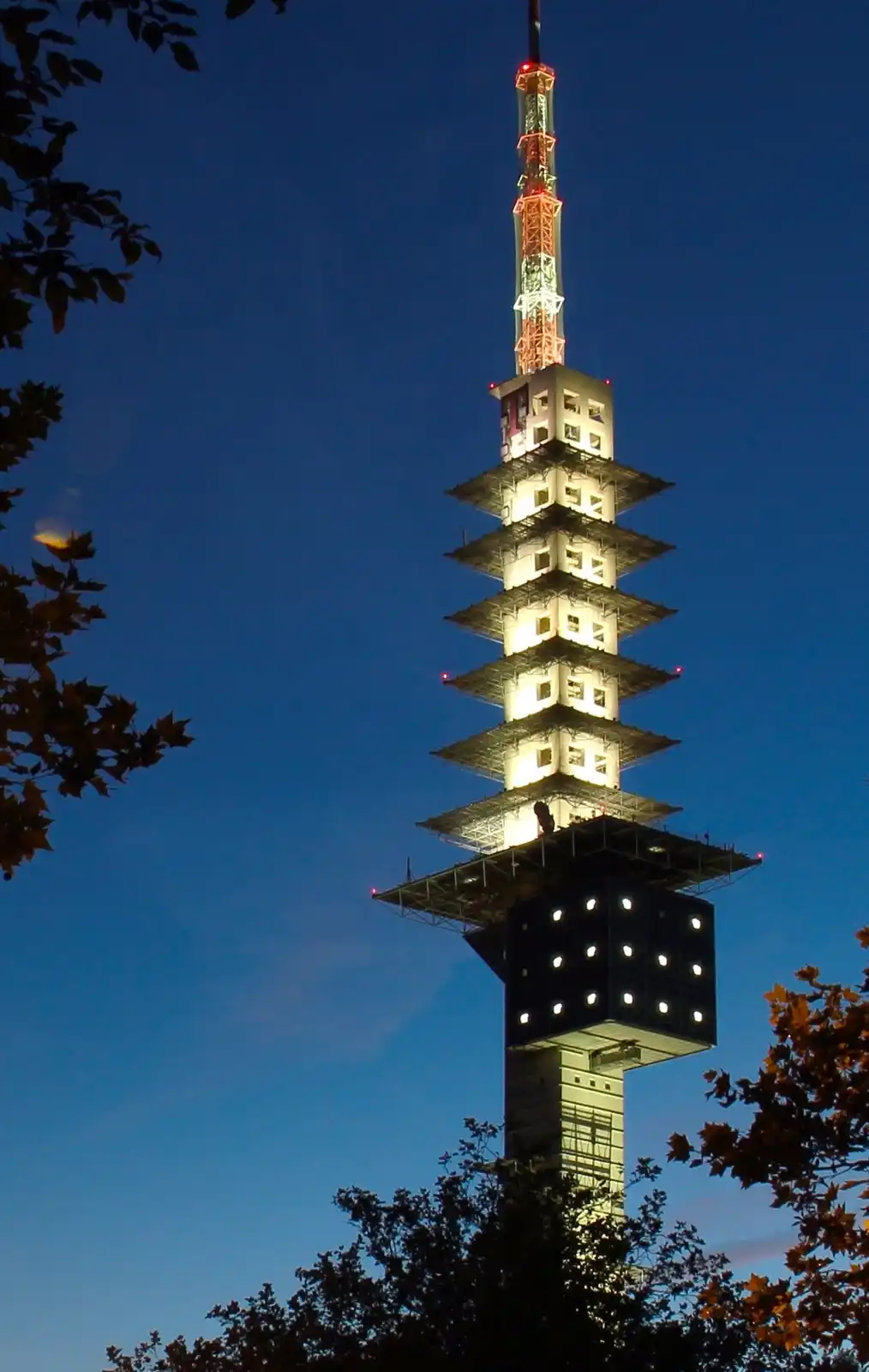 A tall, illuminated tower with a distinctive tiered, pagoda-like upper structure against a twilight sky, framed by tree…