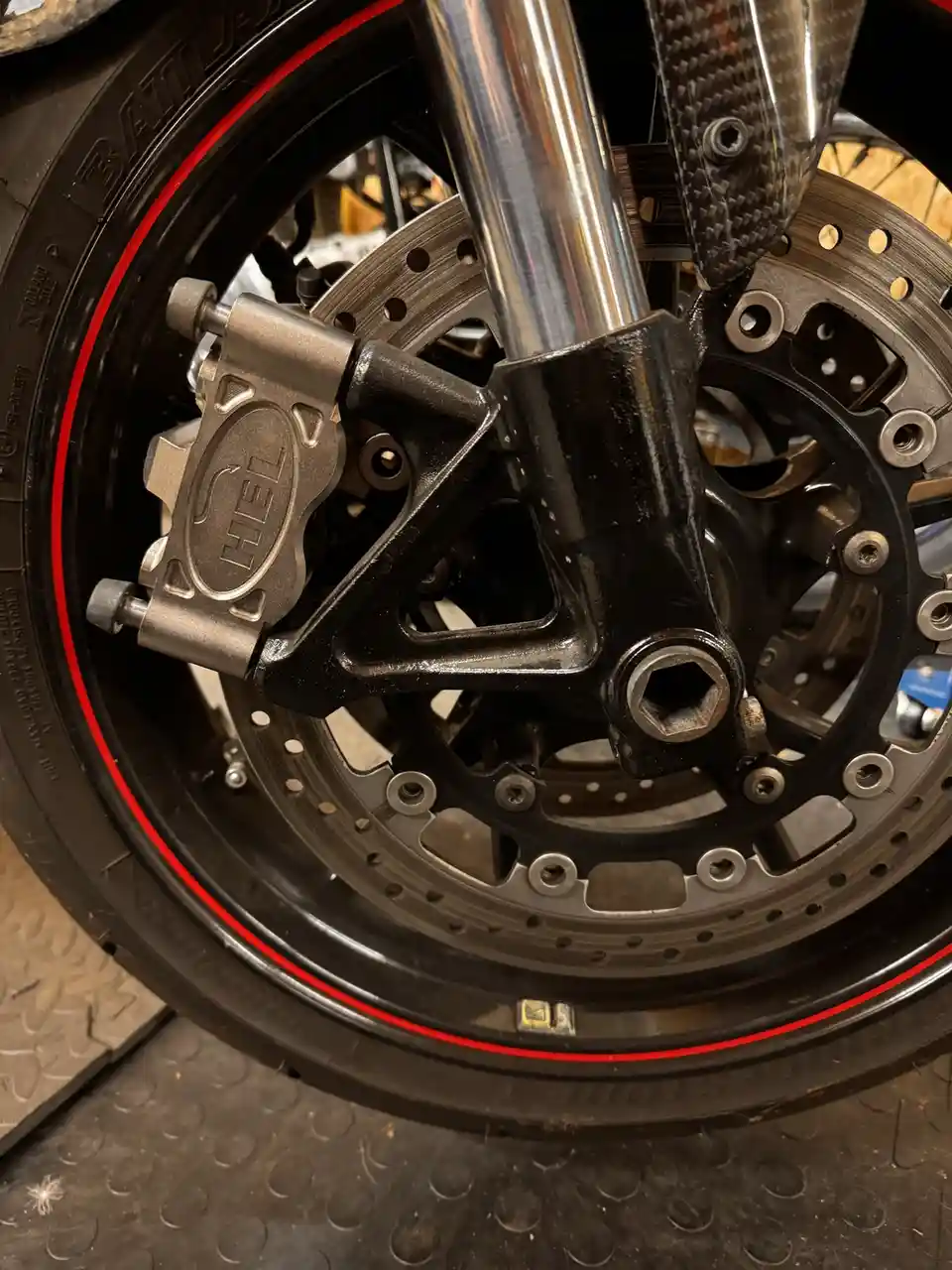 A close-up photo of a motorcycle's front wheel, showing a silver HEL brand brake caliper mounted on a black fork, with a red…