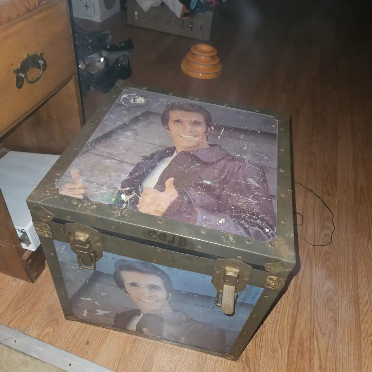 A weathered, olive-green metal trunk with brass latches sits on a wooden floor. The lid and front panel are covered in…