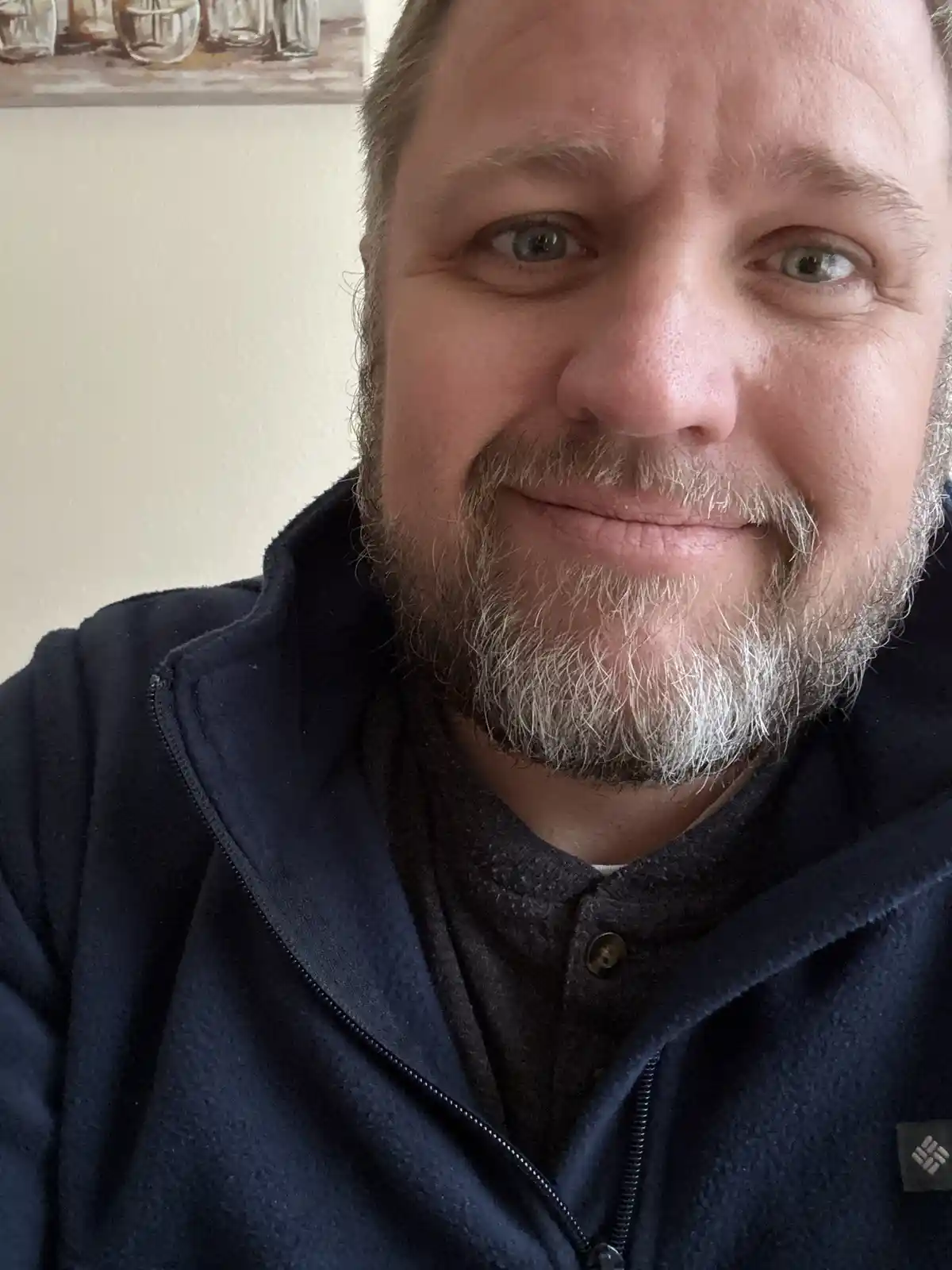 A close-up selfie of a middle-aged man with a salt-and-pepper beard and mustache, wearing a navy blue fleece jacket over a…