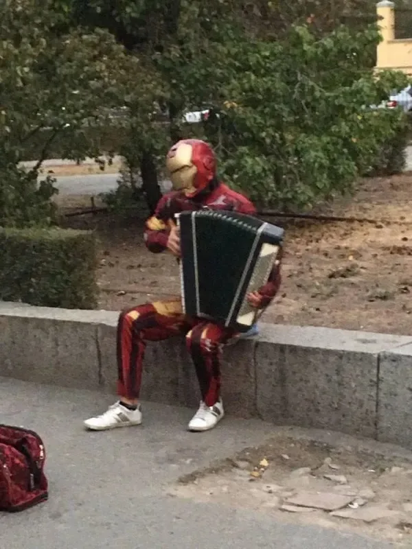 A person dressed in a full Iron Man costume, including helmet and suit, is sitting on a low stone wall outdoors and playing…