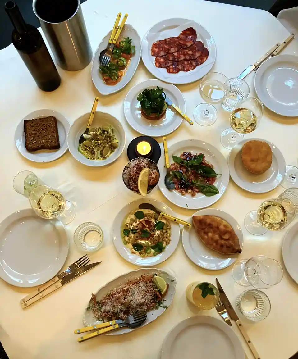 An overhead photo of a white table covered with numerous small plates of diverse gourmet dishes, wine glasses, and cutlery…