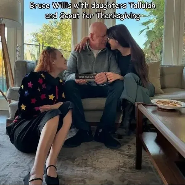 A man, identified as Bruce Willis, sits on a couch holding a plaque that reads 'BEST DAD EVER' while two young women, his…