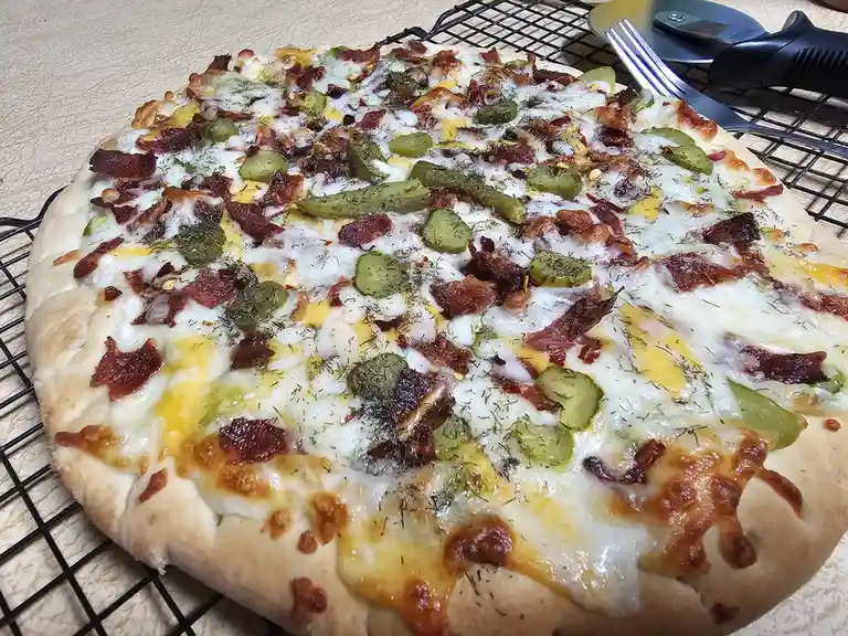 A freshly baked pizza with melted cheese, crispy bacon bits, and green pickle slices sits on a black wire cooling rack. A…