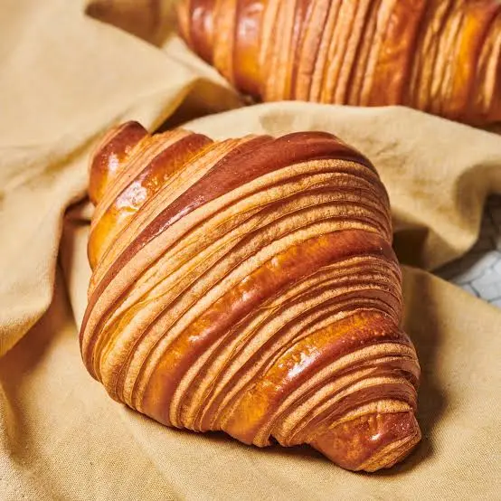 A close-up photograph of a golden-brown, flaky croissant resting on a beige cloth, with another croissant partially visible…