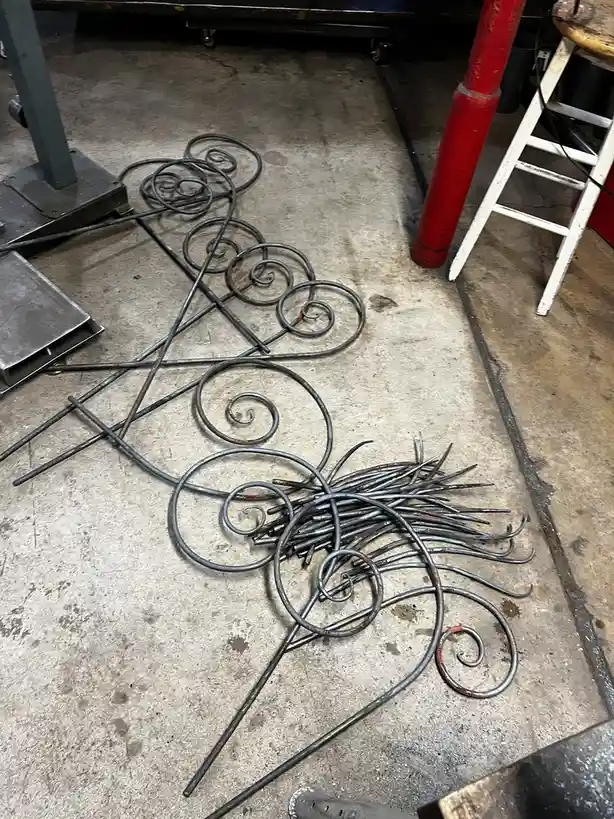 A pile of ornate, scrolled metalwork lies on a concrete floor in a workshop, with a red pole and white stool nearby. The…