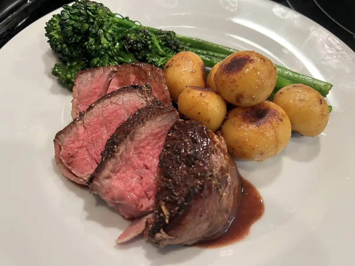 A plate of food featuring sliced medium-rare beef with a dark crust, roasted baby potatoes with charred spots, and steamed…