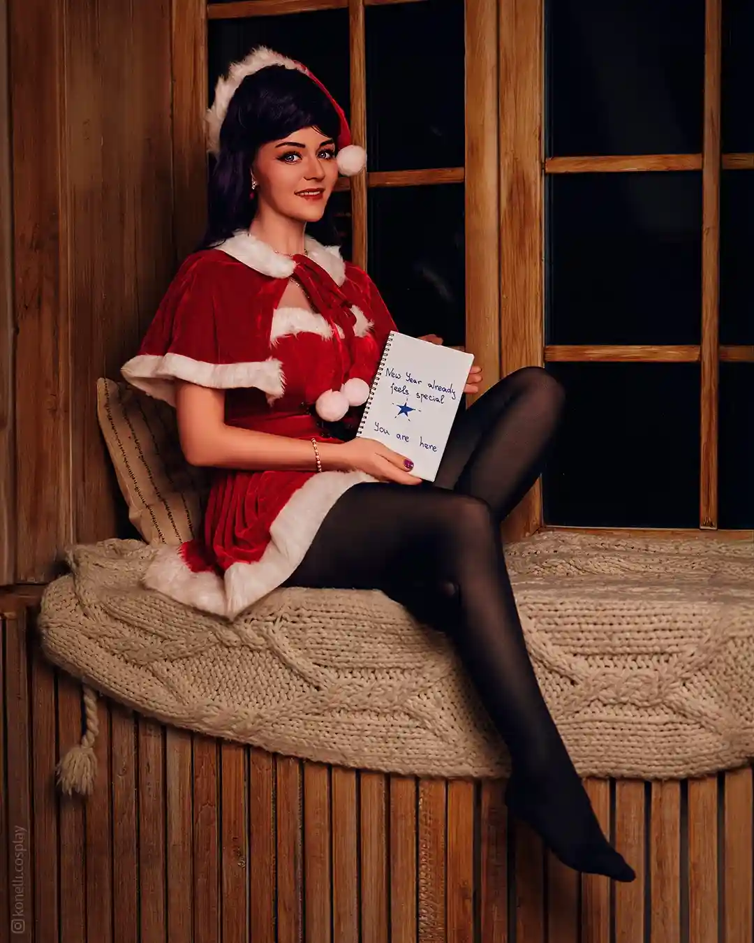 A woman with dark hair wearing a red and white Santa-style outfit sits on a wooden windowsill covered with a thick knit…