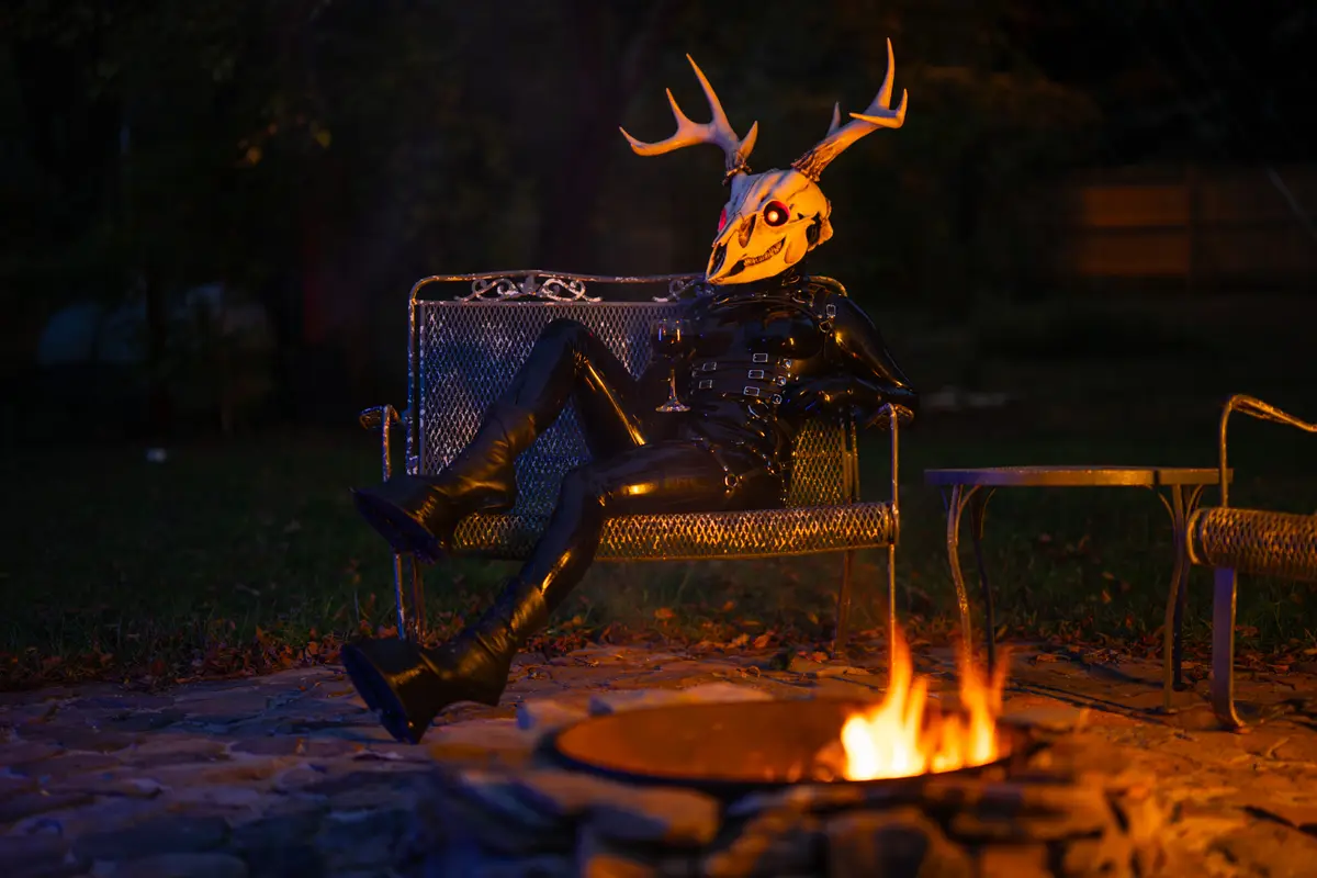 A person in a full-body black latex outfit with a deer skull mask featuring glowing red eyes and antlers is lounging on a…